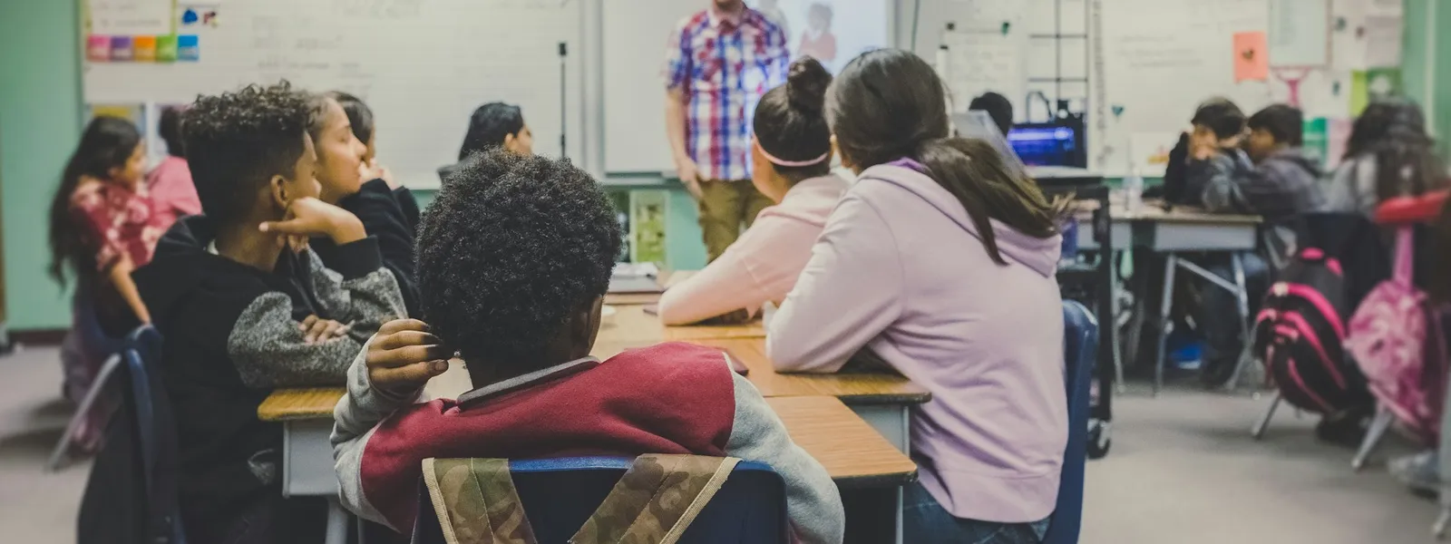 Sala de aula com alunos e professor ao fundo em uma lousa com projetor digital